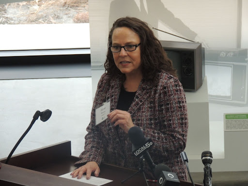 A white woman with brown hair and glasses speaks at a podium.