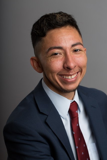 A headshot of Ozzy Orozco, who has brown hair and is wearing a dark blue suit with a red tie. 