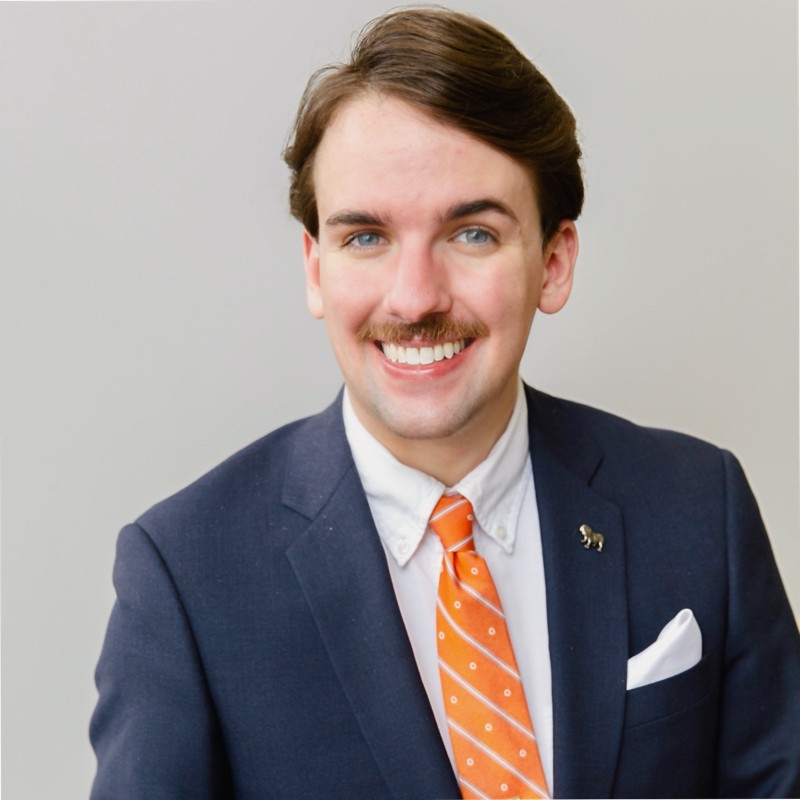 A headshot of Damian Thompson, who is wearing a dark suit with an orange tie. Damian has brown hair and a mustache.