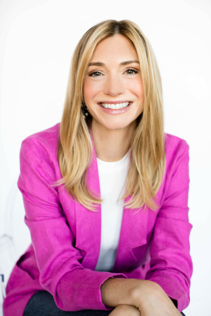 A headshot of Hannah Krause. Hannah has blonde hair and is wearing a bright pink jacket.